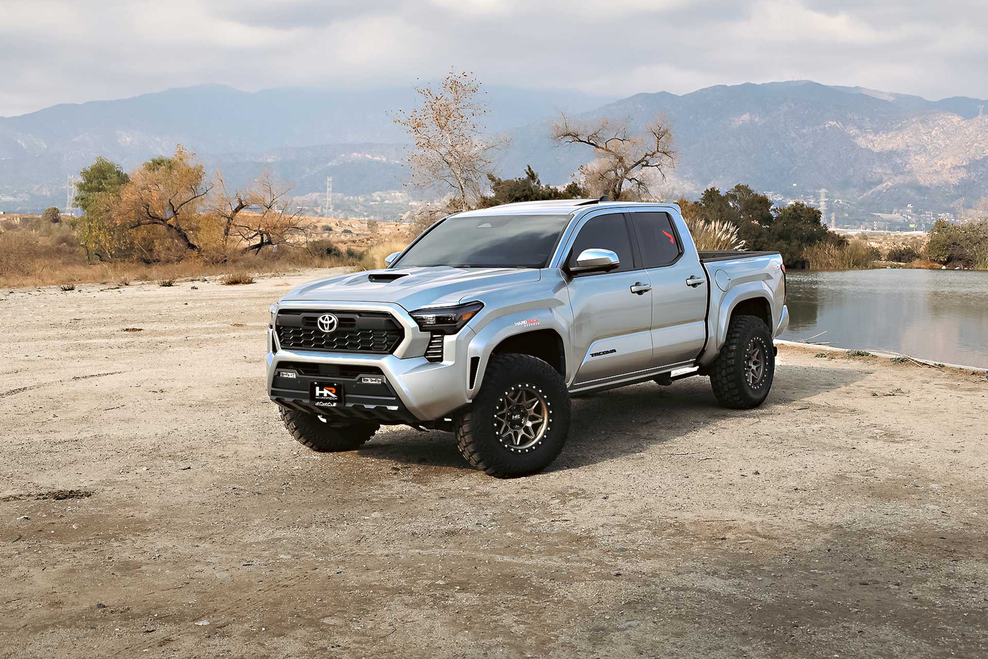 silver gen 4 toyota tacoma trd sport with matte bronze hardrock offroad h105 wheels on dirt by a lake