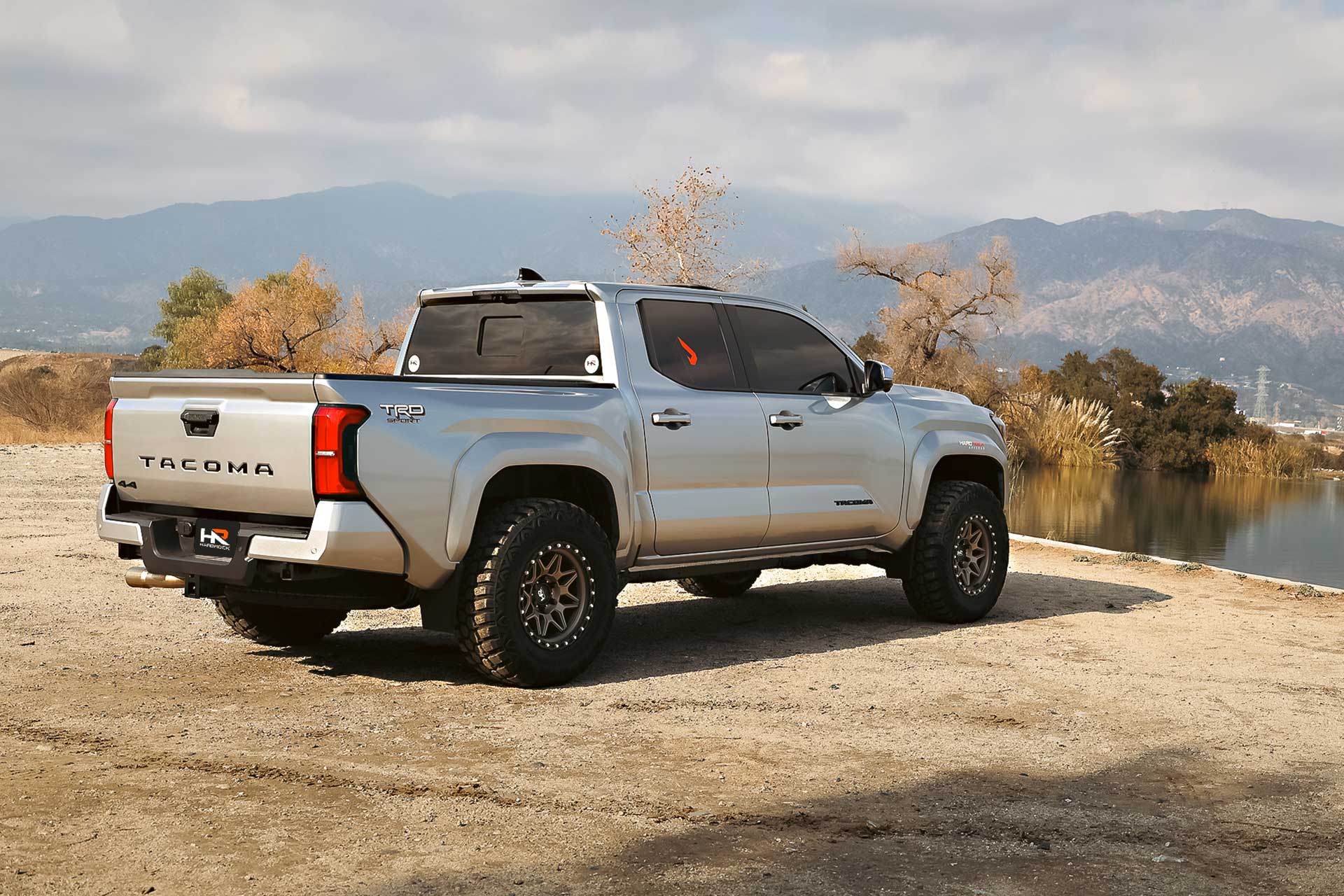 silver gen 4 toyota tacoma trd sport with matte bronze hardrock offroad h105 wheels on dirt by a lake